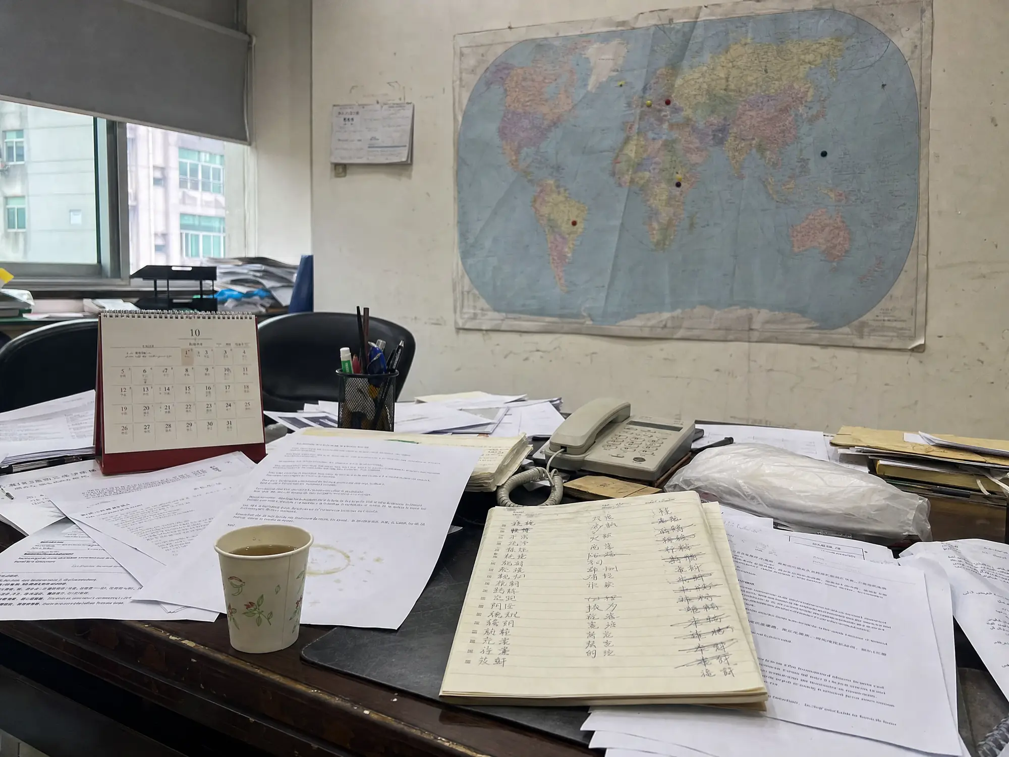 A world map on the wall of a Chinese factory sales office with a few pins stuck in it at random, above a desk buried in printed inquiries from different countries.