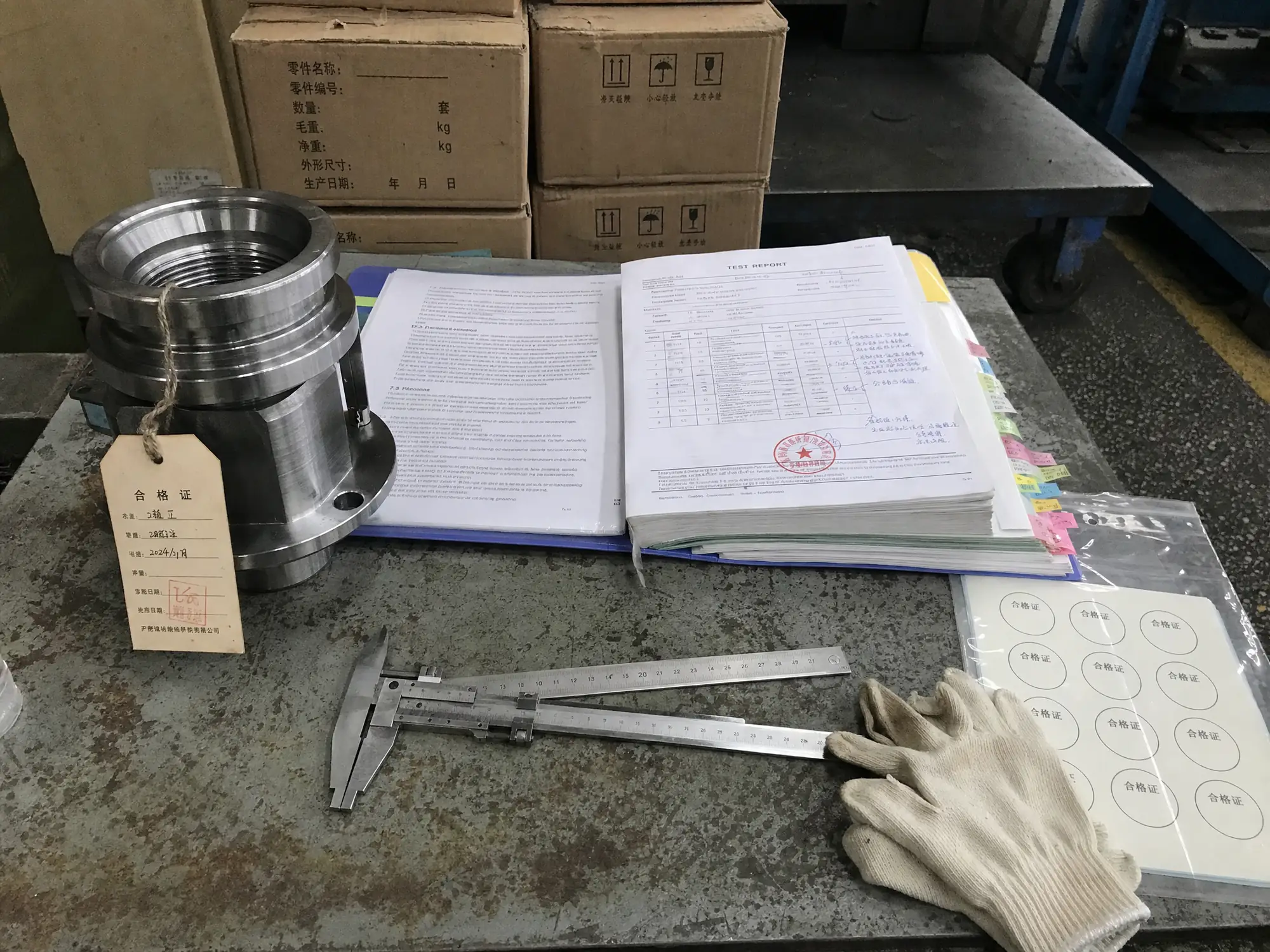 A finished component on a Chinese factory inspection bench beside an open foreign standards binder, a stamped test report, and a sealed sheet of unused certification stickers.