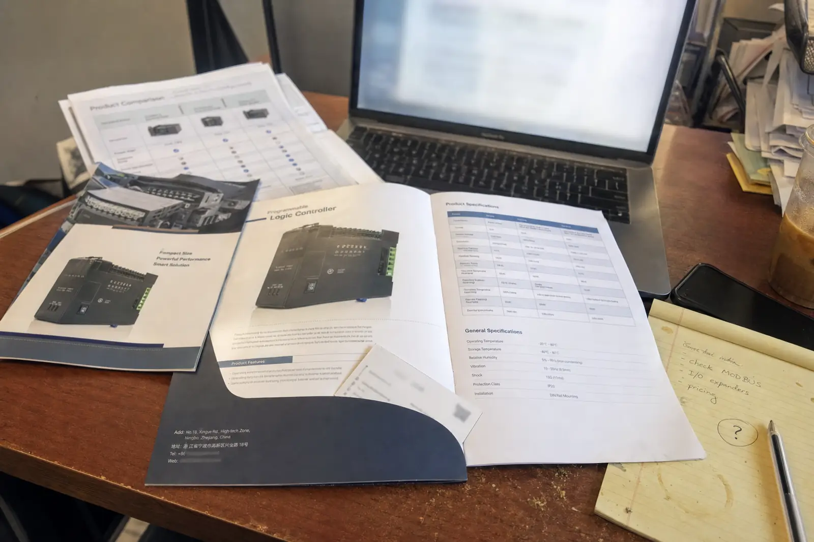A Western buyer’s desk with a Chinese manufacturer’s English-language brochure and specification sheet open on it, the company’s Ningbo address printed on the back cover, and the buyer’s own questions on a notepad.