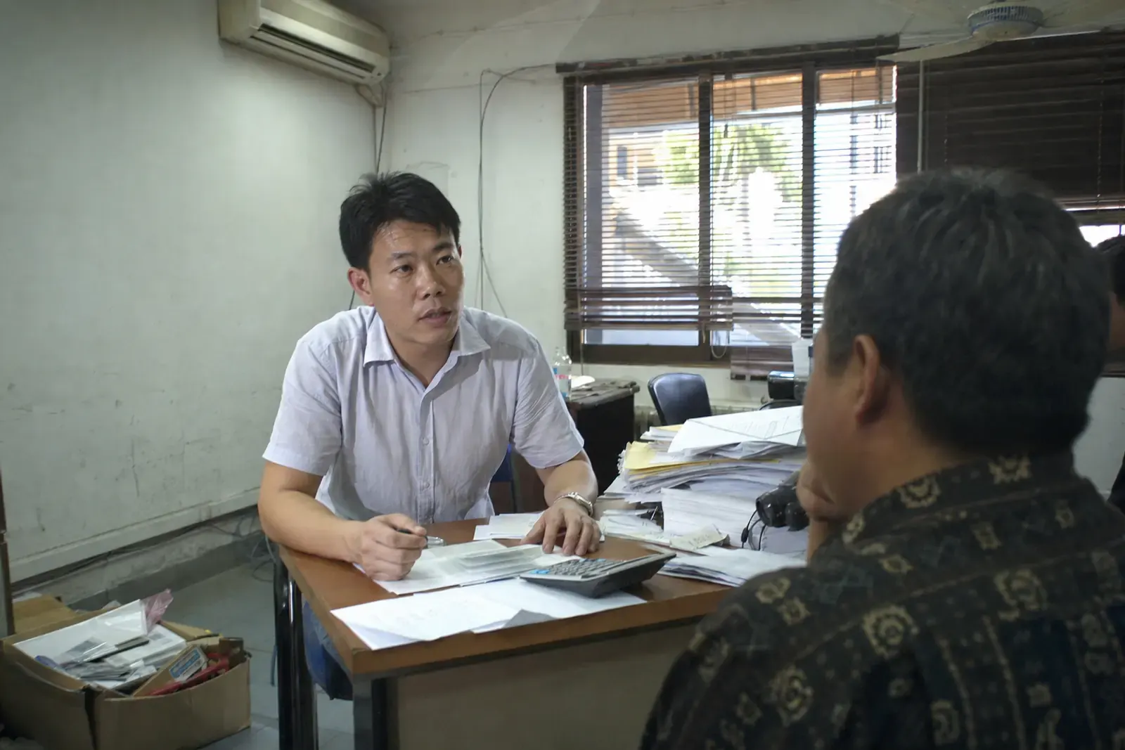 A Chinese export manager meeting a Southeast Asian import agent across a paper-covered desk in a small trading office
